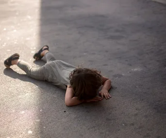 Child lying face-down on pavement, wearing sandals and grey outfit, partially in shadow.