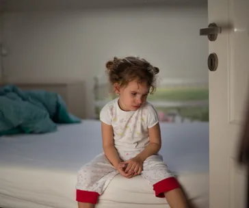Toddler in pajamas sitting thoughtfully on the edge of a bed in a softly lit bedroom.