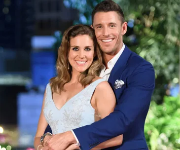 A smiling couple embraces in an elegant garden setting, the woman in a light blue dress and the man in a blue suit.