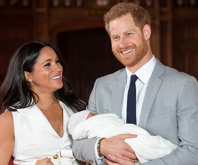 "Couple smiling, holding a newborn wrapped in white, in a formal setting."