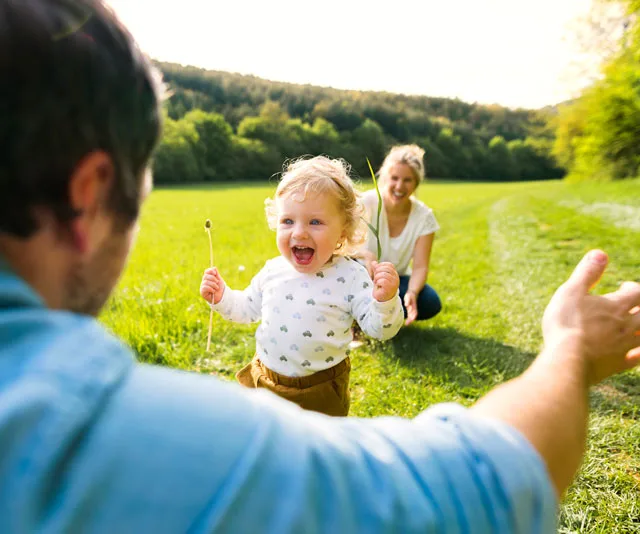 Toddler joyfully running towards an adult in a grassy field, with another adult smiling in the background.