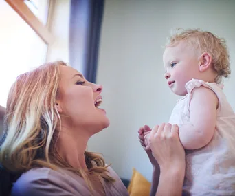 Mother smiling and talking to a 14-month-old toddler, holding hands, near a window.