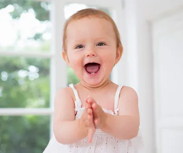 Toddler in a white dress clapping, smiling joyfully in a bright room.
