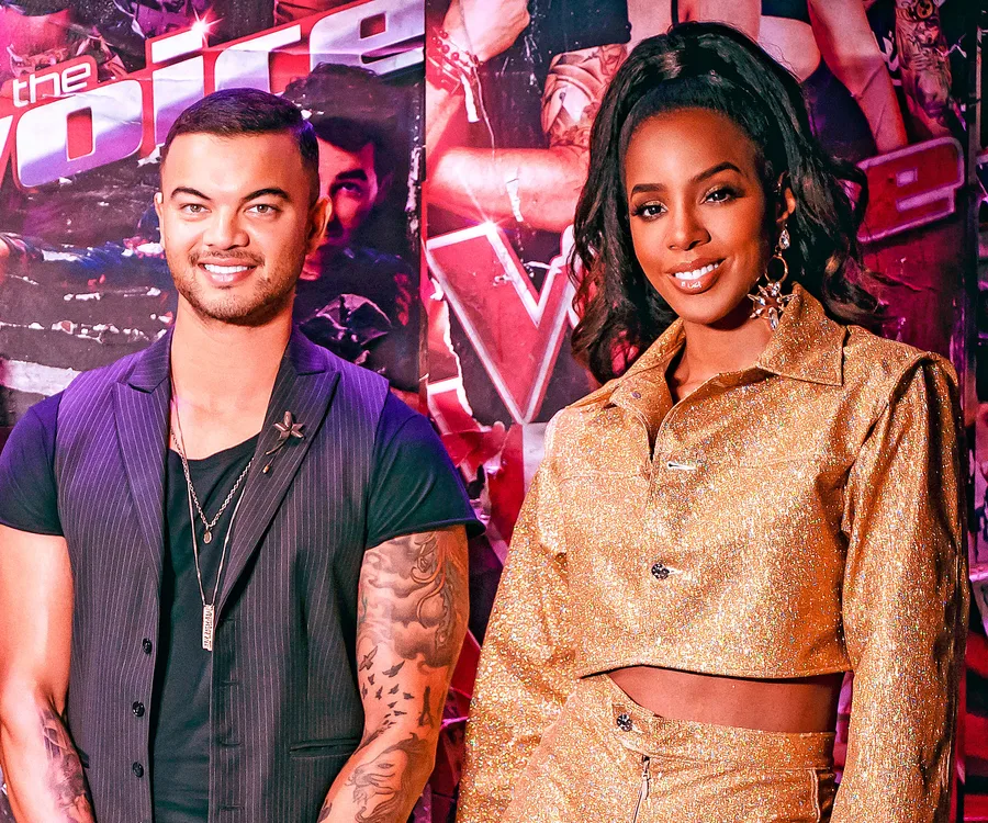 Smiling man and woman in front of "The Voice" backdrop, man in black vest, woman in glittery gold outfit.
