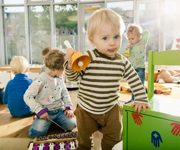 Toddler in striped shirt holds a yellow bell toy in a playroom with other children in the background.