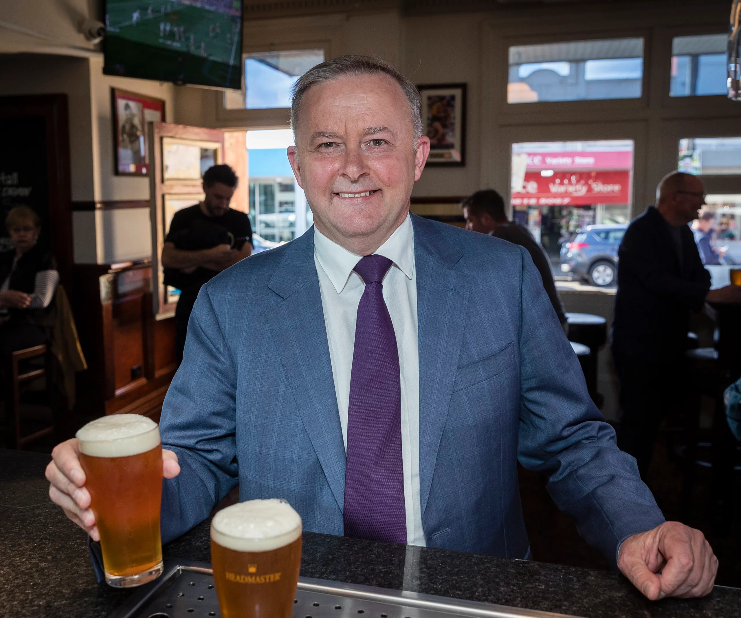 Man in a suit holding a beer at a pub, smiling, with people and a TV showing sports in the background.