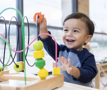A toddler happily plays with a bead maze toy, developing motor skills while sitting by a window.