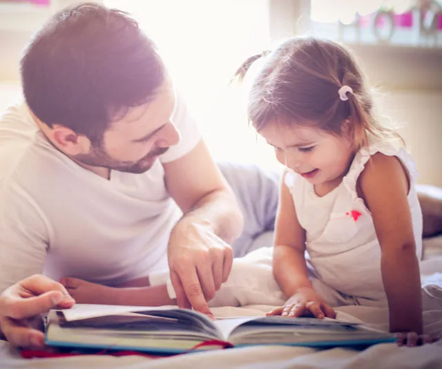 Father and daughter reading a book together on a bed in a warmly lit room.