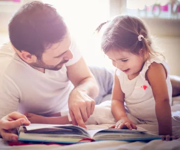 Father and daughter reading a book together on a bed in a warmly lit room.
