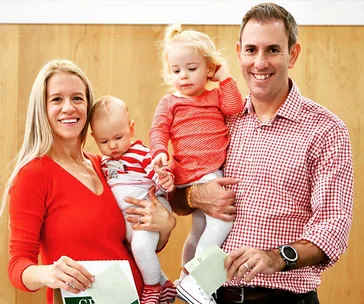A smiling family of four in red attire, holding papers. Two adults and two small children are in a casual setting.