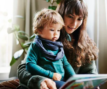 Mother and toddler reading a book together at home, sitting comfortably with plants in the background.