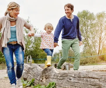 Family walking on a large log outdoors, parents holding child's hands, sunny day, trees in background.