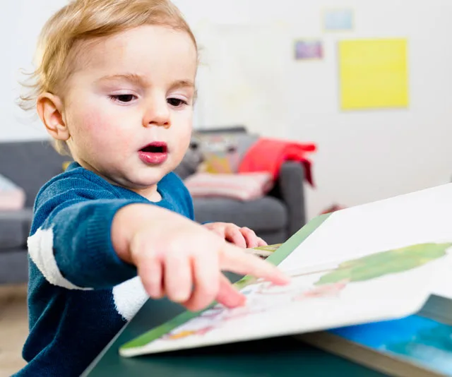 A toddler is pointing at a picture in a book while sitting at a table.
