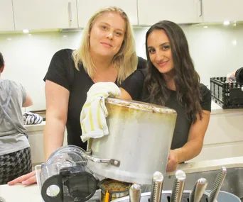 Two women in a kitchen smiling while holding a pot with utensils around.