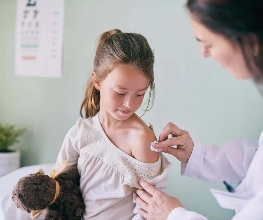 Doctor preparing young girl with a teddy bear for a flu shot, gently cleaning her shoulder.