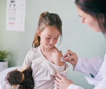 Doctor preparing young girl with a teddy bear for a flu shot, gently cleaning her shoulder.