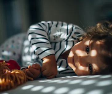 Toddler lying on bed in striped pajamas, holding a toy, with natural light patterns on face.