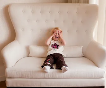 Toddler sitting on a large white armchair covering their face with hands, appearing distressed.