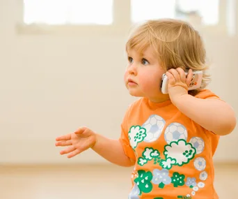 Toddler in orange shirt holding a phone to ear, looking curious.