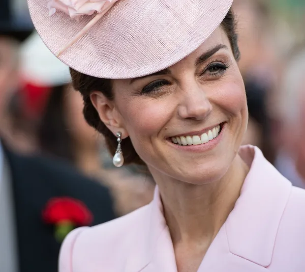 A smiling woman in a pink outfit and hat, wearing elegant pearl drop earrings.