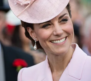 A smiling woman in a pink outfit and hat, wearing elegant pearl drop earrings.