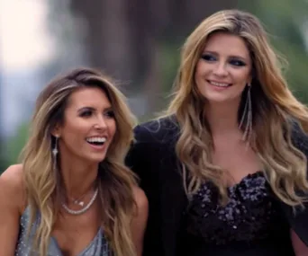 Two smiling women with long hair walk side by side in elegant attire outdoors.