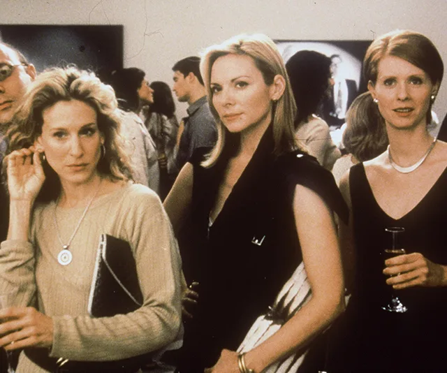 Three women at a social gathering, each holding a drink, with a crowd in the background.