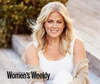 Blonde woman smiling, sitting outdoors on steps, wearing a white top and beige cardigan, with magazine logo at bottom left.