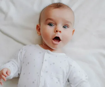 A surprised baby in a white onesie with stars, lying on a soft white surface.