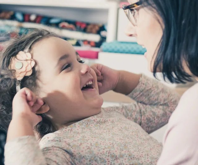 Child with flower headband laughing and holding ears, interacting with adult, conveying a joyful moment together.