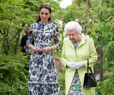 Royal figures walking together in a garden, one in a floral dress, the other in a light green coat.