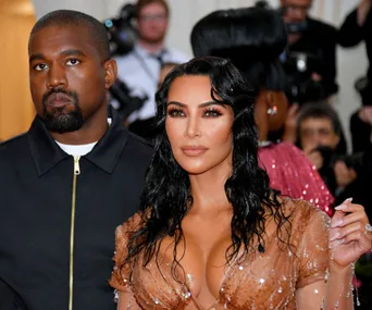 A man in a dark jacket and a woman in a sheer, embellished gown attending a public event, facing the camera.