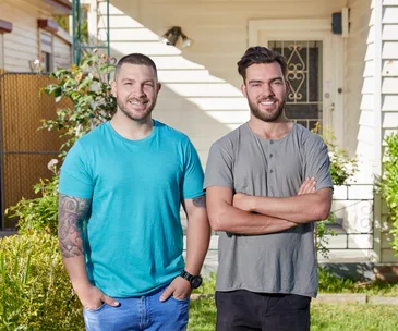 Two men standing outside a house. One with a tattooed arm and wearing a blue shirt, the other in a gray shirt.