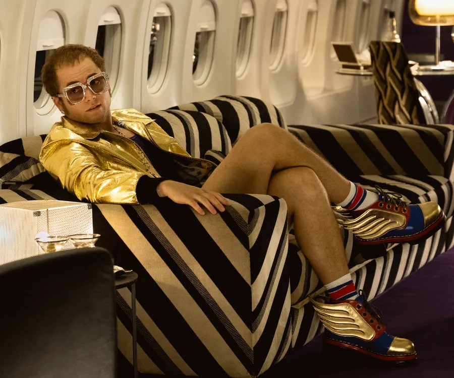 A person in a golden jacket and sunglasses lounges on a striped sofa inside an airplane, wearing colorful winged shoes.