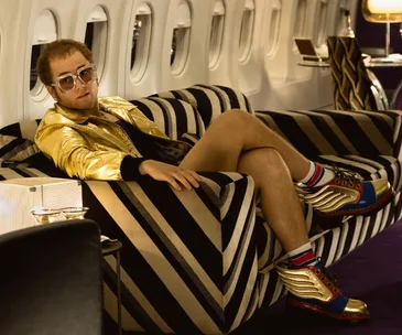 A person in a golden jacket and sunglasses lounges on a striped sofa inside an airplane, wearing colorful winged shoes.