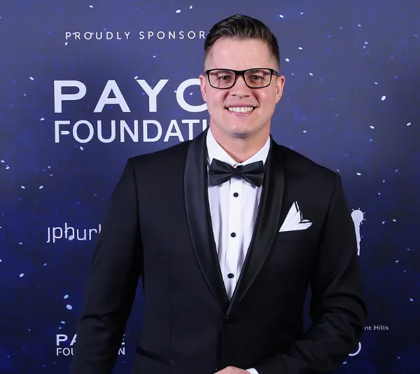 Man in tuxedo with glasses smiling at a gala event, standing against a blue "PAYCE Foundation" backdrop.