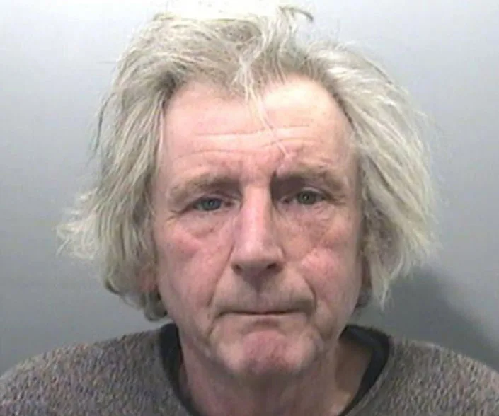 Elderly man with tousled gray hair and a solemn expression against a plain background.
