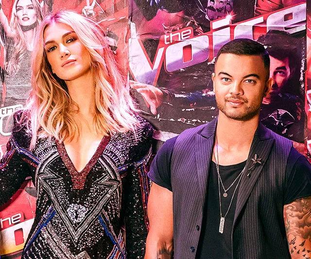 Pair of judges pose in front of "The Voice" backdrop, dressed stylishly.