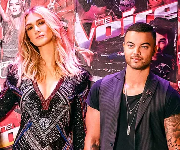 Pair of judges pose in front of "The Voice" backdrop, dressed stylishly.
