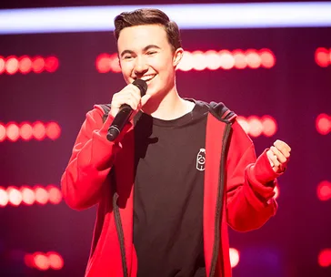 A person sings on stage in a red jacket, holding a microphone, with red lights in the background.