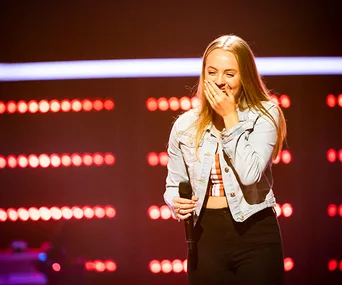Contestant on stage holding a microphone, smiling with hand over mouth, wearing a denim jacket and checkered top.