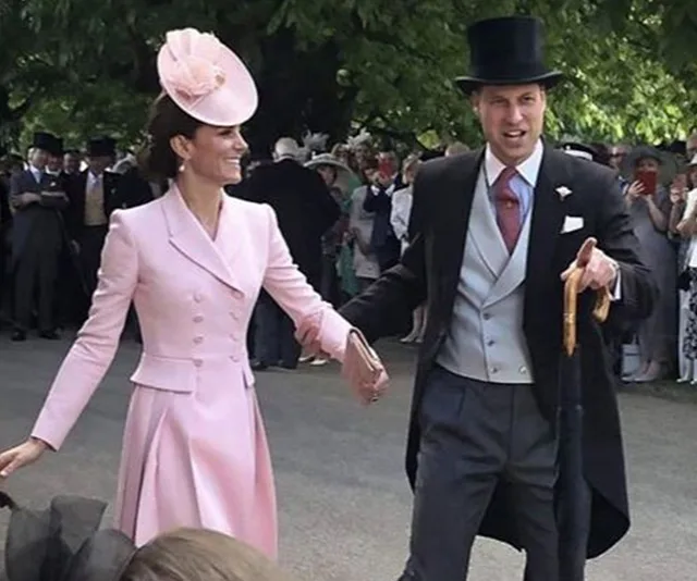 A couple elegantly dressed, holding hands and walking outdoors, surrounded by a crowd.