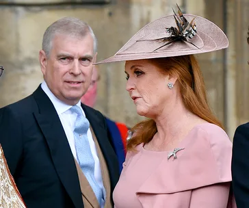 Two individuals in formal attire at an outdoor event, with one wearing a pink dress and hat adorned with feathers.