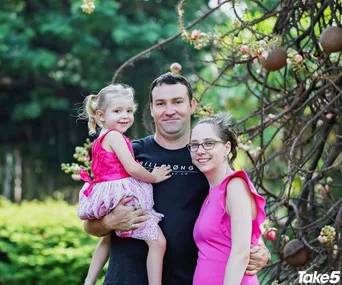 A family of three in a garden, with a man holding a young girl, and a woman standing beside them, all smiling.