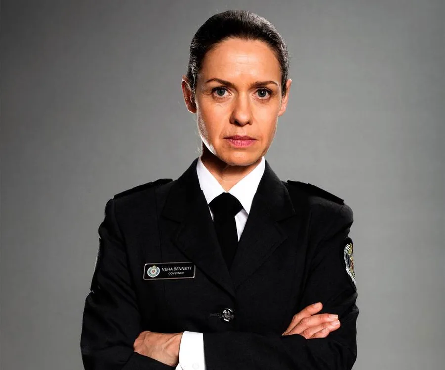 Vera Bennett from Wentworth in a black uniform, arms crossed, serious expression against a grey background.