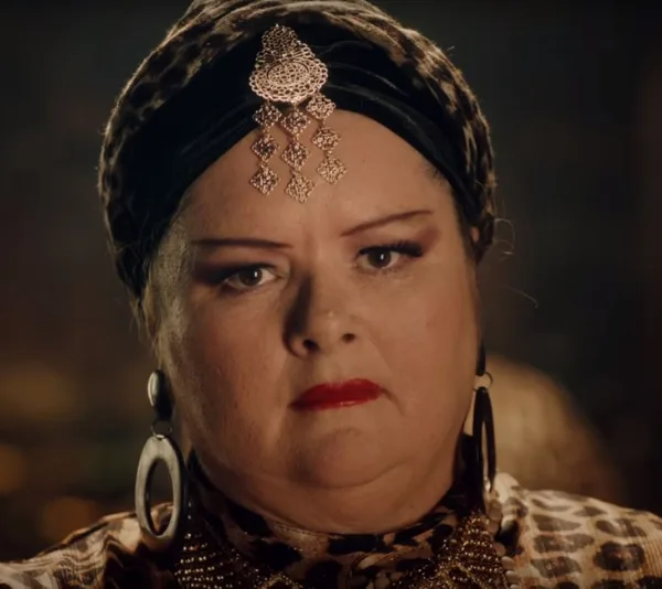 A person wearing a leopard print headdress, large earrings, and a decorative headpiece, with a serious expression.