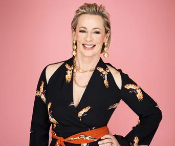 A woman in a black dress with bird designs and statement jewelry, smiling against a pink background.