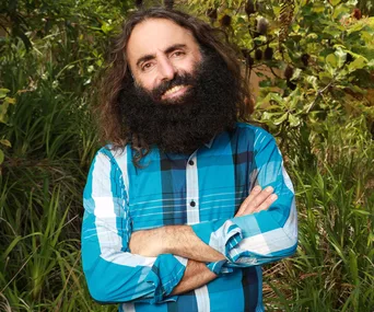 Man with long beard and blue plaid shirt stands smiling in a lush garden.