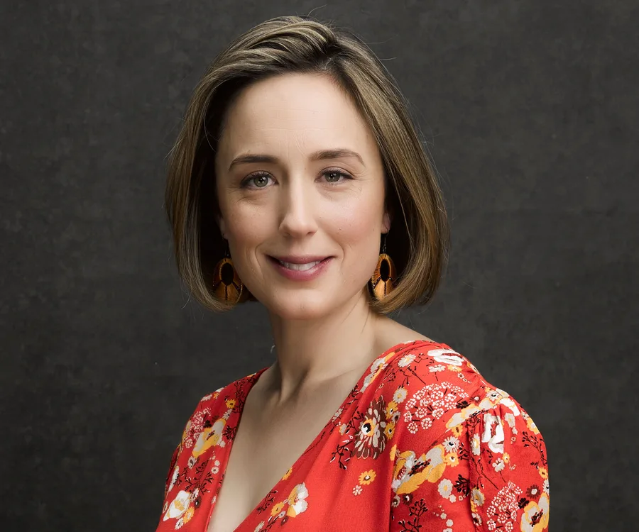 A woman wearing a red floral dress and orange earrings smiles against a dark background.