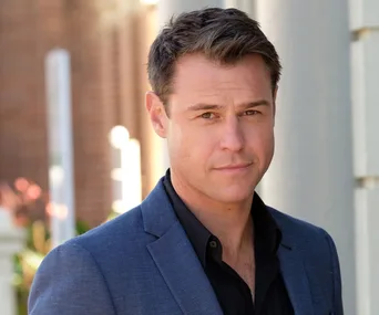 Man in a blue suit and black shirt, outdoors near a brick wall; neutral expression.